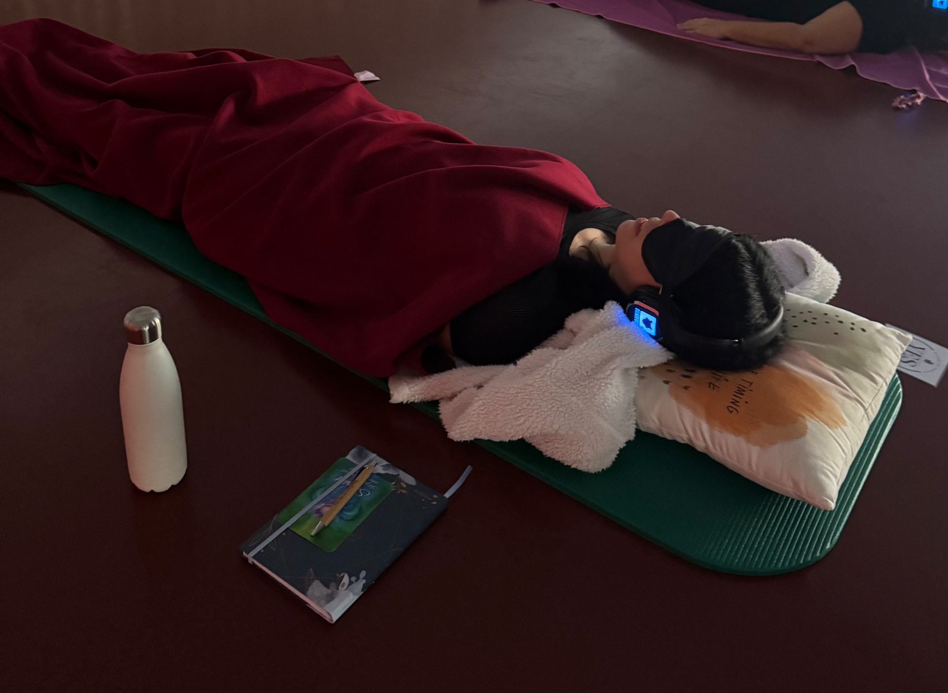 A participant rests on a mat during a breathwork session, wearing headphones and an eye mask, covered with a blanket.